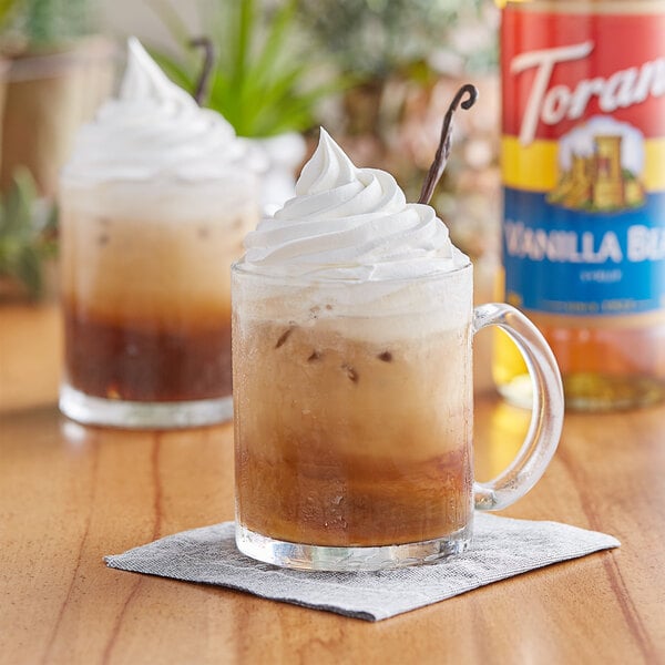 A glass mug of brown liquid with a Torani Vanilla Bean drink and whipped cream on a table.