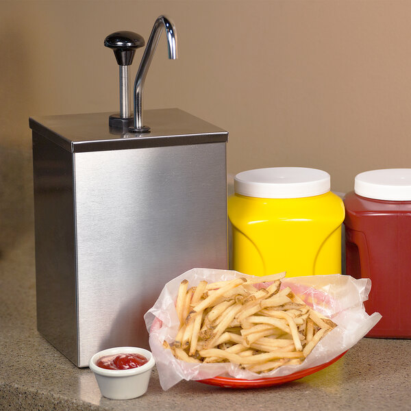 A stainless steel Carlisle high volume condiment pump over a yellow container of mustard on a counter with french fries and ketchup.