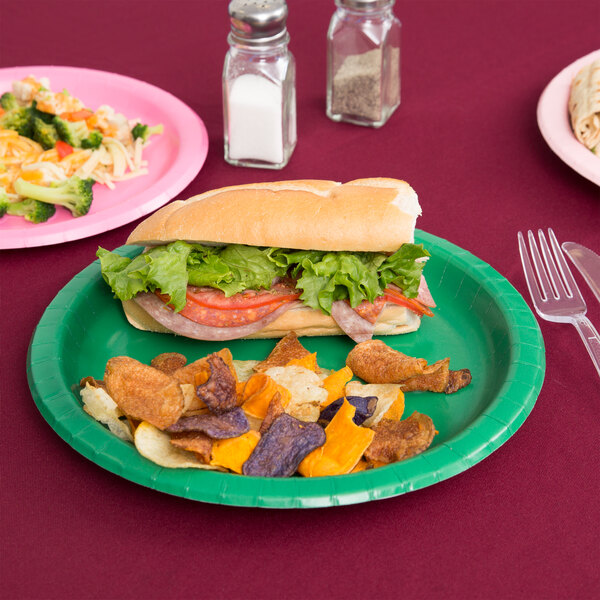 An emerald green Creative Converting paper plate with a sandwich, lettuce, tomato, and meat on a table.