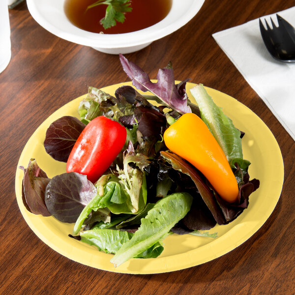A Creative Converting mimosa yellow paper plate with a salad, small pepper, and lettuce on it on a table with a bowl of soup and a fork.
