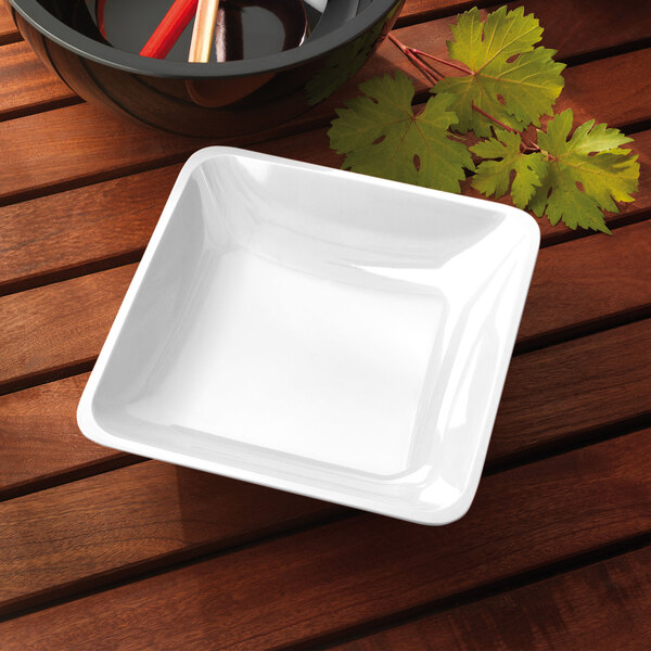 A white square Elite Global Solutions melamine bowl on a wooden table.