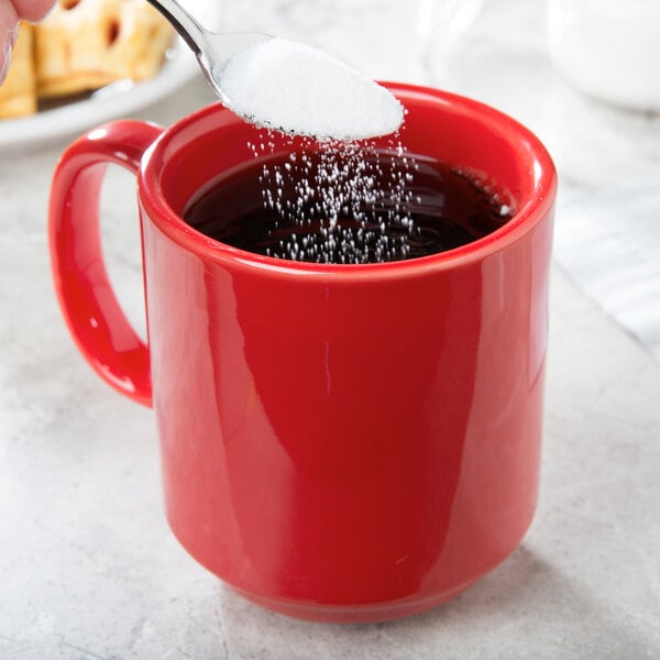A spoonful of white sugar being poured into a red CAC Venice mug.