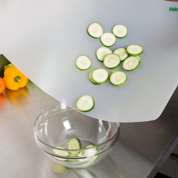 A person using a WebstaurantStore flexible cutting board to slice a cucumber over a bowl of peppers.