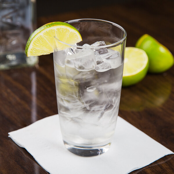 A highball glass of water with ice and lime wedges on a table.
