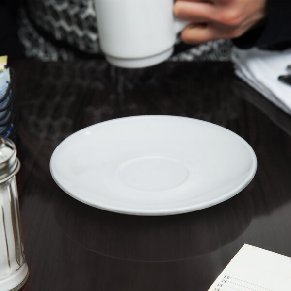 A white saucer on a table with a white Arcoroc cup on it.