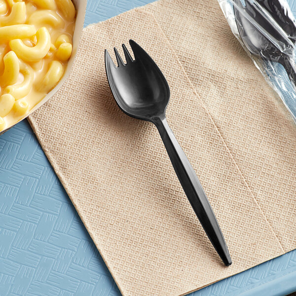 A black plastic spork on a napkin next to macaroni and cheese.
