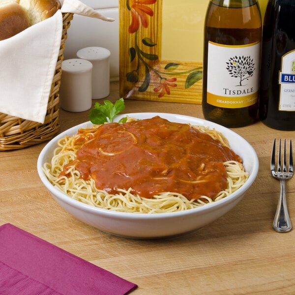 A white Tuxton pasta bowl filled with spaghetti and sauce on a table with wine bottles.