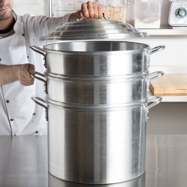 A man in a white chef's coat opening a large aluminum clam steamer pot.