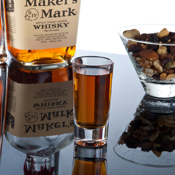 A Thunder Group plastic dessert shot glass filled with brown liquid next to a bottle of liquor and a bowl of nuts.