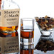 A bottle of whiskey next to an Arcoroc shot glass filled with brown liquid and a bowl of nuts.