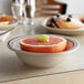 A brown speckle stoneware bowl with a grapefruit slice in it on a table with a plate of food.