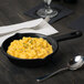 A Tablecraft black and green speckled cast aluminum fry pan of macaroni and cheese on a table with a fork and spoon.