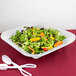 A salad with lettuce, carrots, and a pair of white scissors in a white square bowl.