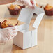 A person in gloves opening a white Baker's Mark cupcake box with a muffin inside.