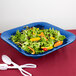 A Tablecraft cobalt blue cast aluminum square bowl filled with salad and oranges on a blue plate with spoons.