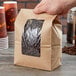 A hand holding a brown Kraft paper bag with a window filled with coffee beans.