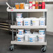 A gray Metro utility cart with three shelves holding white and clear plastic containers of food.