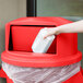 A hand putting a white container into a red Rubbermaid trash can.