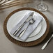 A white plate with a chestnut rattan charger plate on it with silverware and a white napkin.