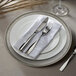 A white plate with a silver rim on a white napkin with silverware.