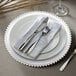 A white Charge It by Jay glass charger plate with clear beaded rim on a table set with silverware and a napkin.