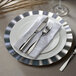 A silver plastic charger plate with silverware on a white napkin.