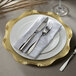 A white plastic charger plate with a baroque gold rim, silverware, and a napkin on a table.