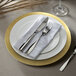 A white Charge It by Jay plastic charger plate with a gold rim and silverware on it.