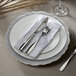 A white plate with a silver scalloped design on the edge with a silver fork and knife on a white napkin.