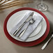 A white plastic charger plate with a red rim on a white background with silverware and a napkin.