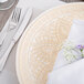 A white glass charger plate with a gold pattern on it, set on a table with a napkin and silverware.
