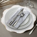 A white Charge It by Jay Baroque plastic charger plate with silverware and a napkin on it.