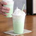 A hand holding a plastic cup with Big Train Dragonfly Green Tea Blended Creme Frappe Mix in a green drink with whipped cream.