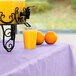 A glass of orange juice sits on a lavender plastic tablecover on a table.