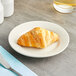 An Acopa ivory wide rim stoneware plate with a croissant on it next to a knife and fork.