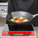 A Vollrath carbon steel non-stick fry pan with a black and silver handle on a stove with meat and vegetables cooking in it.