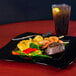 A Fineline black plastic square plate with steak, shrimp, and vegetables on a table with a drink.