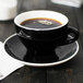 A black CAC Venice cup with a white saucer on a table.
