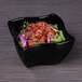 A black Elite Global Solutions square melamine bowl filled with food on a table.