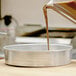 A person pouring brown liquid into an American Metalcraft aluminum round cake pan.