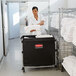 A woman in a white shirt pushing a black and silver Rubbermaid laundry cart full of towels.