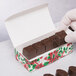 A gloved hand holding a Poinsettia holiday candy box with a row of chocolates inside.