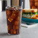 A 32 oz. clear plastic tumbler filled with ice tea on a table with a sandwich.