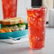 A clear plastic tumbler filled with red liquid and ice on a table with a sandwich in the background.