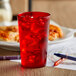 A red GET plastic tumbler filled with red liquid and ice on a table with a plate of pizza.