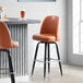 A Lancaster Table & Seating caramel brown vinyl bar stool with swivel bucket seat next to a table.