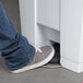A person's foot in jeans and sneakers stepping on the pedal of a white rectangular Rubbermaid commercial trash can.
