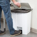 A person in jeans putting a cup into a white Rubbermaid step-on trash can.