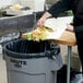A person cutting food on a counter and throwing it into a Rubbermaid Brute trash can.