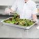 A woman in a white shirt and gloves holding a Western Plastics foil steam table pan filled with salad.